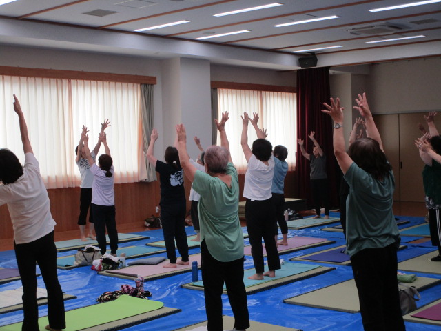 フローリングの広い室内で、青いビニールシートの上にヨガマットを敷いた多くの参加者が、立ち上がって両手を上に伸ばしてストレッチを行っている写真
