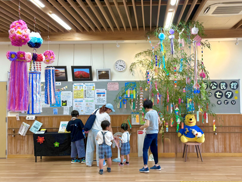 明るい室内の広間に、天井から吊り下げられた大きな飾りと、短冊やカラフルな飾りが付けられた七夕の笹飾りが立てられており、数人の親子が笹の周りで飾り付けや掲示物を見ている様子の写真