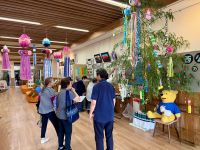 天井から吊るされた七夕飾りと、短冊やカラフルな飾りが賑やかに飾られた大きな笹の葉が目を引く室内で、数人の高齢者たちが集まって展示物や笹飾りを眺めている様子の写真