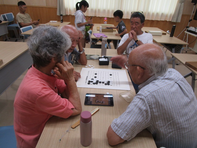 明るい室内で、4人の高齢者がテーブルを囲んでおり、中央に置かれた碁盤で真剣に碁石を打ちながら、その横のタブレットも利用して囲碁の対局を楽しんでいる様子の写真