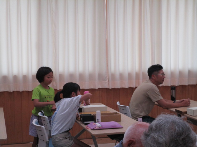 明るい室内で、白いカーテンがかかった窓を背に、2人の幼い女の子が小さな碁盤のようなものを使って遊んでおり、その奥では一人の男性が座って様子を見ている様子の写真