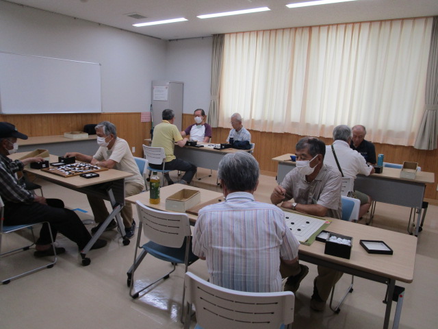 明るい室内で、数台の長机に高齢の男性たちがそれぞれ向かい合って座り、マスクを着用しながら碁石や碁盤を使い真剣に囲碁の対局を楽しんでいる様子の写真