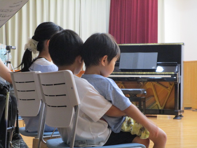 背景には黒いアップライトピアノと赤いカーテンが見える室内で、白い椅子に男の子が幼い男の子を抱っこして座っており、その隣にはポニーテールの女の子が座っているイベントの様子の写真