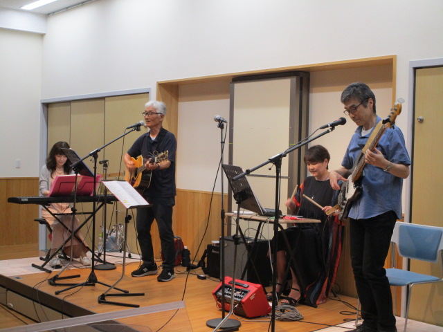 ステージの上で4人組のバンドが演奏しており、左からキーボードの女性、アコースティックギターとボーカルの男性、電子パーカッションの女性、ベースギターの男性がそれぞれの楽器を演奏しているライブ写真