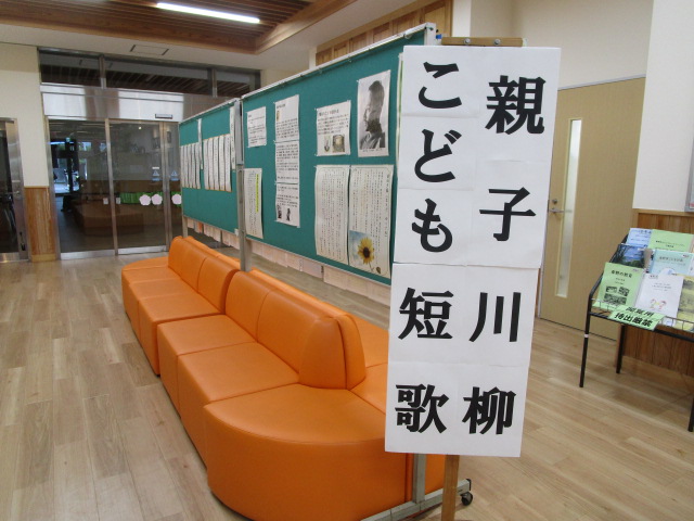 木目調の床と明るい室内空間に、オレンジ色の長いソファと、緑色の掲示板が設置されており、その手前には「親子短歌川柳」と縦書きされた大きな看板が置かれている写真