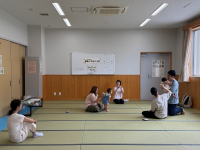 明るい畳敷きの広い部屋で、数組の親子が床に座っており、中央でつかまり立ちをしている赤ちゃんを囲んで親たちが手遊びのような交流を楽しんでいる様子の写真