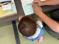 畳の上に立っている赤ちゃんがテーブルの上に手を置いており、その横で大人の手が緑色の用紙にスタンプを押すかサインをしている様子を上から捉えた写真