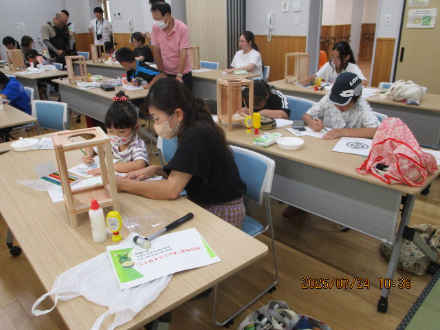 明るい室内で、木製の枠組みのあんどんのようなものを囲んで、子供と大人がテーブルに向かい、色を塗ったり絵を描いたりする工作活動に熱中している様子の写真