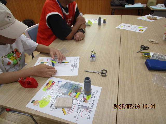 明るい室内で、ベージュのテーブルに広げられた資料を見ながら、キャップをかぶった子供が小さなモーターや電池を使った工作に取り組んでおり、隣には指導者と思われる大人が座っている科学工作教室の様子の写真