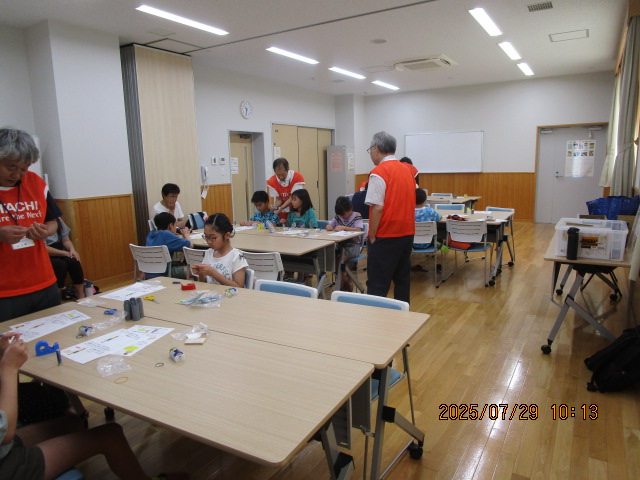 明るいフローリングの室内で、数台の長机を囲んで座っている子供たちが、ゴムや紙などの材料を使い工作に取り組んでおり、オレンジ色のベストを着た大人が指導している科学工作教室の様子の写真
