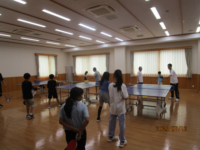 フローリングの広い室内で、複数の卓球台にラケットを持った子供たちが並んで対面にいる大人と卓球の練習をしている写真
