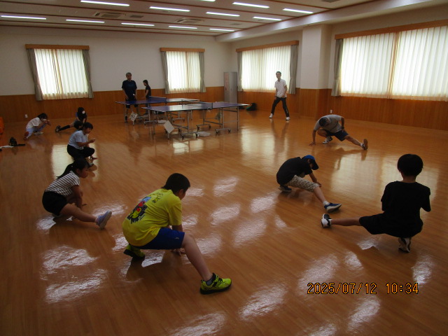 フローリングの広い室内で、卓球台が数台置かれている体育館のような場所で、数人の子供たちが大人と一緒に足を開いて前屈する整理運動を行っている様子の写真