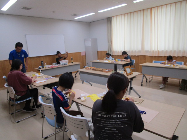 明るい会議室のような部屋で、数台の長机を使い、数組の親子がそれぞれ座って紙やハサミなどの道具を使って熱心に工作に取り組んでおり、一人の男性が指導している様子の写真