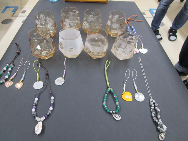 黒いテーブルの上に、サンドブラストで模様が施された琥珀色の八角形のガラスコップが8個並べられており、その手前にビーズやペンダントトップを使った手作りのアクセサリーが多数置かれている写真