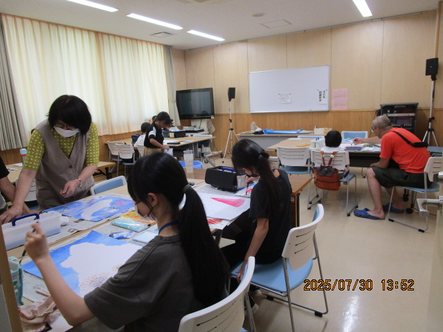 明るい会議室のような部屋で、長机を使い複数の参加者が絵の具で水彩画のような制作活動に取り組んでおり、マスク姿の指導者と思われる女性が手前の参加者にアドバイスをしている様子の写真