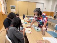 部屋の長机を囲み、カラフルな服を着た年配の女性が、数人の子供たちに、きんちゃく袋作成の作業を指導している様子の写真