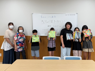 白いボードの前に、2人の大人と4人の子どもが並んで立っており、子どもたちは自分で作ったカラフルな巾着袋を手に持って笑顔でカメラを見ている、手芸教室の記念写真