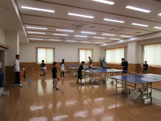 明るいフローリングの広い室内で、複数の卓球台の周りに子供と大人が集まり、それぞれラケットを持って大人が子供に卓球を指導している写真