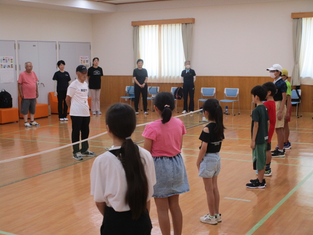 体育館のようなフローリングの部屋で、ネットを挟んで、中央に立つ指導者の男性を中心に、数人の大人と子供たちが整列して話を聞いている様子の写真
