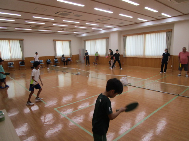 フローリングの広い室内で、ネットを挟んで、数人の子供と大人がパドルを持ち、ラインで区切られたコート内でフリーテニスを楽しんでいる写真