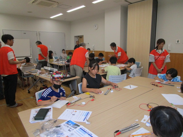 明るい教室のような室内で、数人の子供たちがテーブルでリニアモーターカーの実験キットと思われるものを組み立てており、オレンジ色のベストを着た大人が指導にあたっている科学工作教室の様子の写真