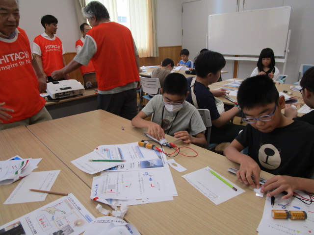 明るい室内で、数人の子供たちがテーブルに広げられた資料を見ながら電池や導線を使って工作に取り組んでおり、後ろではオレンジ色のベストを着た大人が指導している科学工作教室の様子の写真