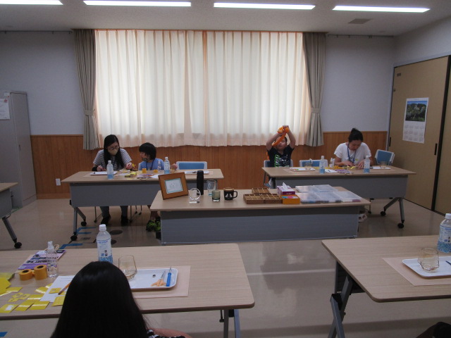 白いカーテンのかかった窓を背にした室内で、長机を囲んで数組の親子が座っており、テーブルの上には工作や学習用の道具が置かれている、サンドプラスト彫刻体験の様子の写真