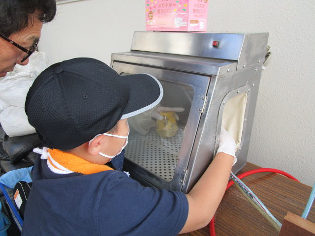 マスクとキャップを着用した少年が、ステンレス製の箱状の機械を手袋をはめ、中にある黄色い物体を操作しており、隣で眼鏡をかけた大人が覗き込んでいる様子の写真