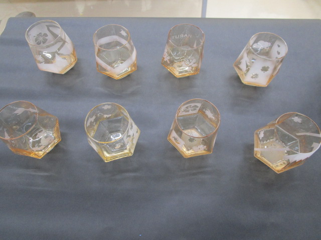 黒い布の上に、桜やクローバーなどの模様がサンドブラストで描かれた、琥珀色をしたガラスのコップが合計8個並べられている様子を上から捉えた写真