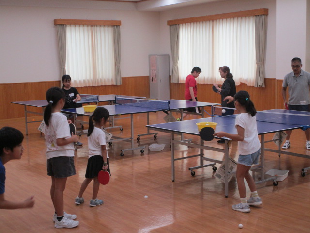 木目調の壁と明るいフローリングの室内で、複数の卓球台が設置されており、子供と大人がラケットを持って卓球の練習を楽しんでいる様子を捉えた写真