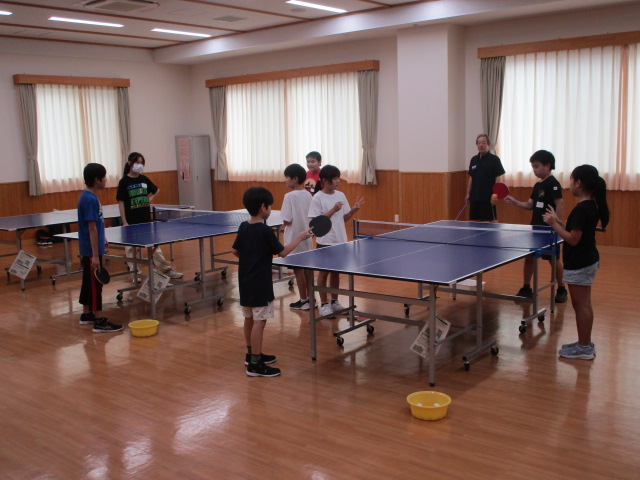 明るいフローリングの室内で複数の卓球台が設置されており、子供たちが卓球のラケットを持ち、大人の指導者がいる中で練習や交流を楽しんでいる様子を捉えた写真