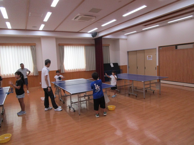 フローリングの明るい室内で複数の卓球台が設置されており、子供たちが試合をしているのを大人が審判をして見ている写真