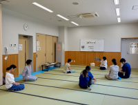 明るい畳敷きの広い部屋で、数人の女性と一人の男性が円形に座って静かに話を聞いており、中央で一人の女性が絵本を持って読み聞かせをしている様子の写真