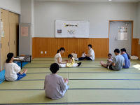 畳敷きの広い部屋で、親と思われる大人たちが子どもたちを連れて車座になって座っており、中央で一人の女性が幼い女の子と手遊びをしている様子の写真