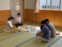 畳敷きの広い部屋で、3組の家族と高齢の女性が一堂に会し、中央に広げられたカードや本を見ながら、床に座って穏やかな交流を楽しんでいる様子の写真