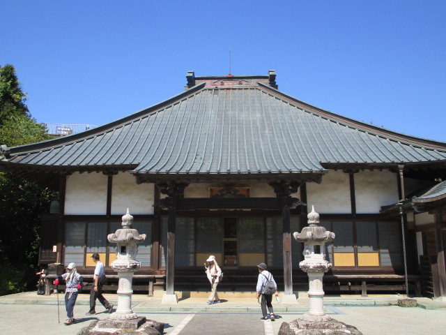 青空の下、数名の観光客や参拝者が重厚な瓦屋根と白い壁を持つ日本の伝統的な仏堂前の石灯籠の間を歩いている様子を正面から捉えた写真