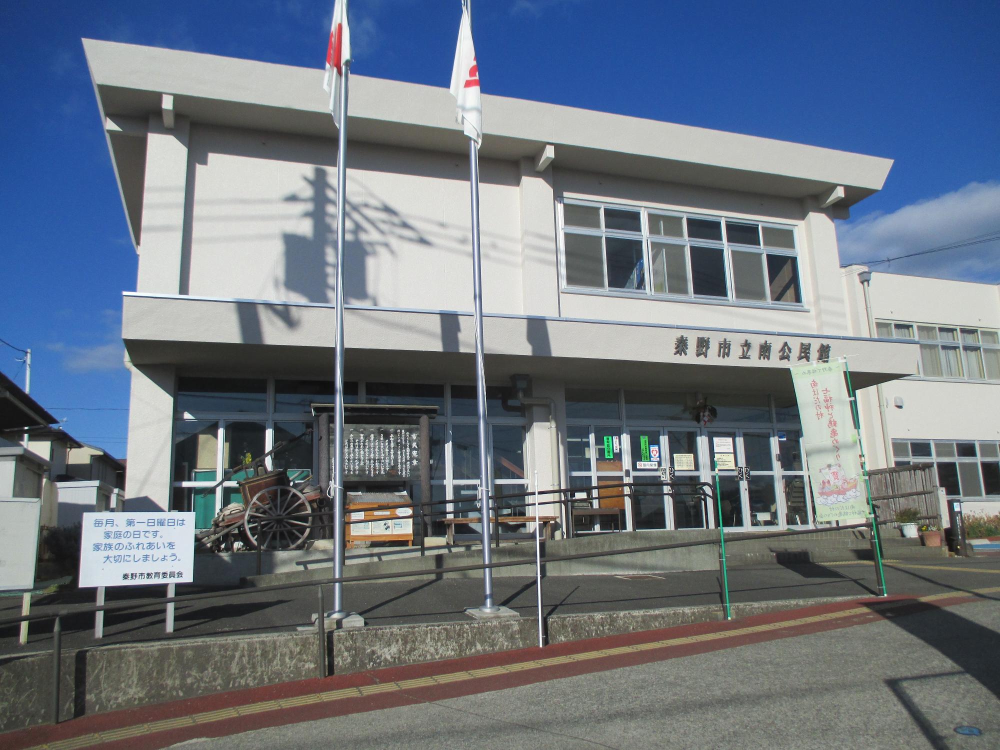 青空のもと二本の旗が掲揚され、入り口付近には木製の荷車と掲示板があり、「東野市立南公民館」と書かれた看板が見えるベージュ色の二階建ての建物の正面を写した写真