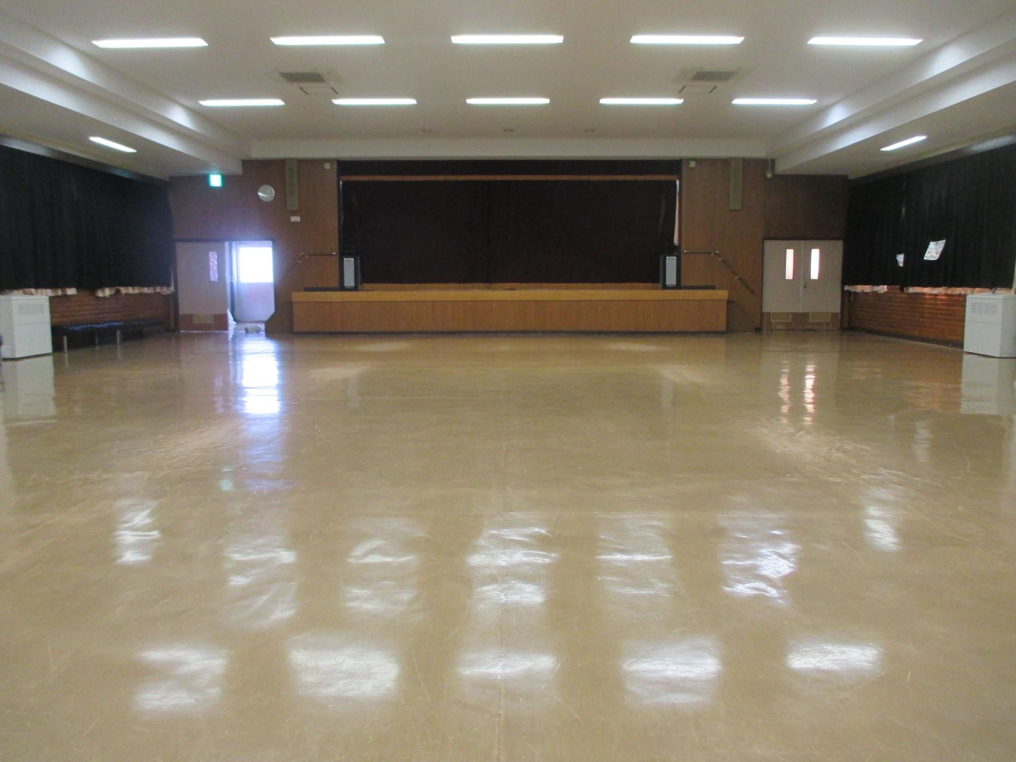 蛍光灯が並ぶ白い天井、磨かれた木製フロア、正面に黒い幕と木製ステージ、両側に木目調の壁面装飾を持つ多目的ホールの写真