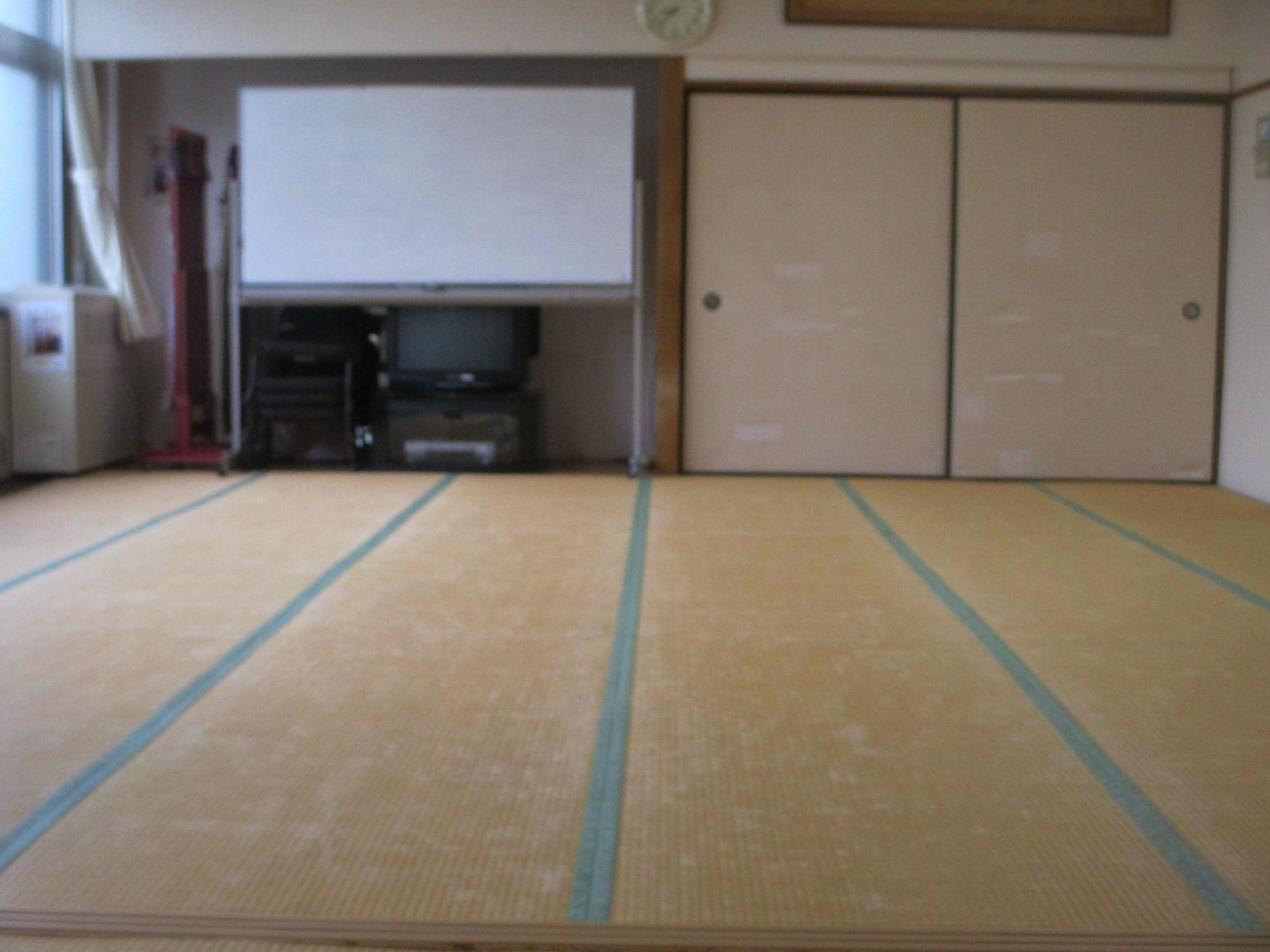 畳が敷かれた和室の室内で、正面に白いスクリーンが設置された収納スペースと薄茶色の襖が並び、左側には窓と椅子が配置された和室27畳の写真