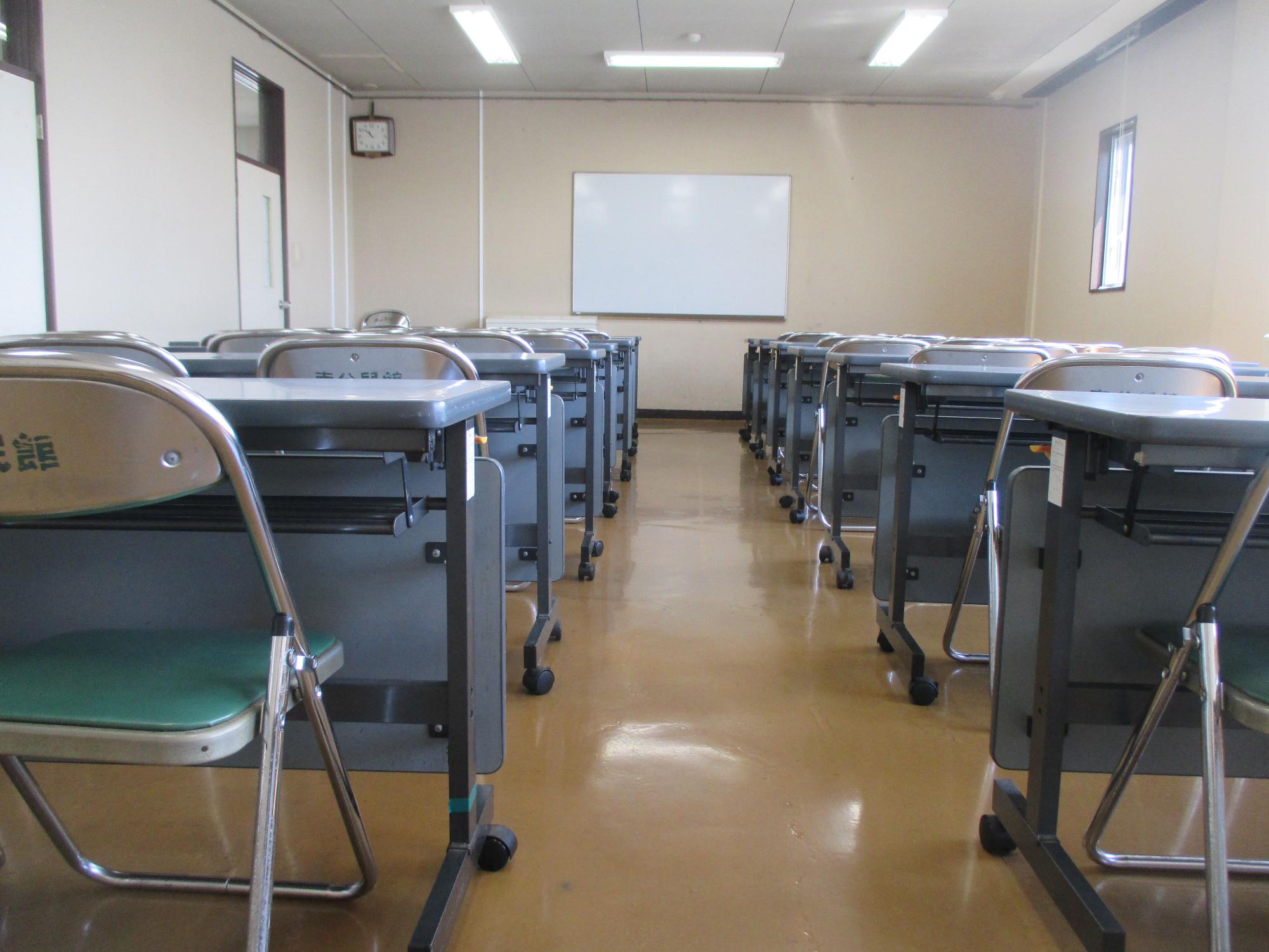 蛍光灯照明のある白い天井、薄茶色の壁に取り付けられた白いホワイトボード、そして緑色の座面を持つ折りたたみ椅子とグレーの机が整然と並ぶ集会室の写真