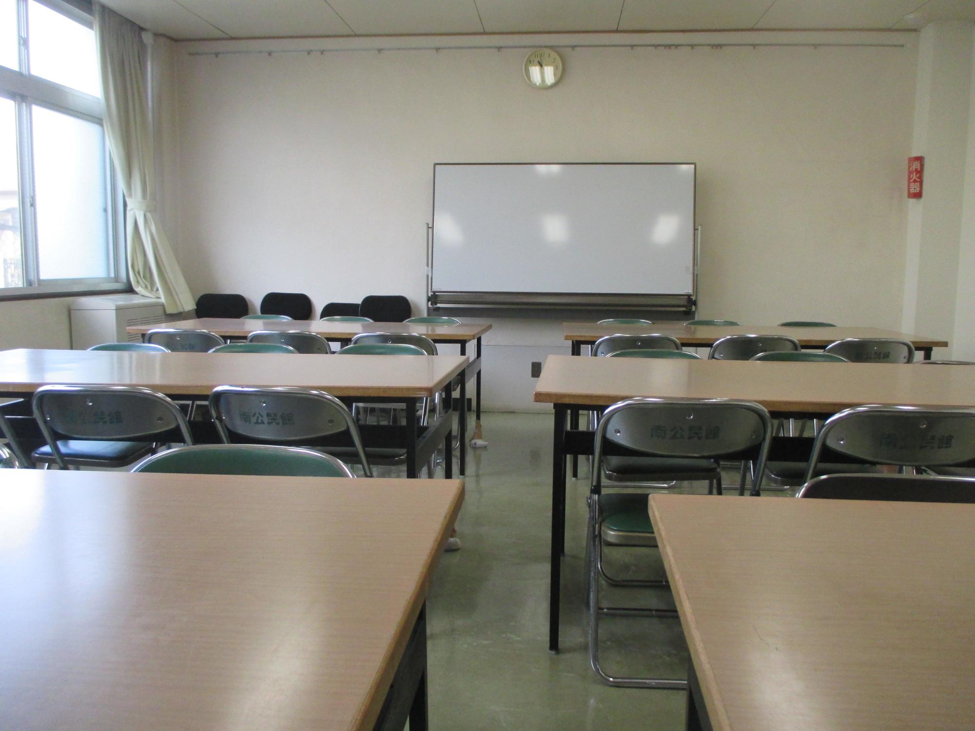 左側に大きな窓とカーテン、正面中央に白いホワイトボードと時計、そして緑色の座面を持つ折りたたみ椅子と木製天板の机が整然と配置された学習室の写真