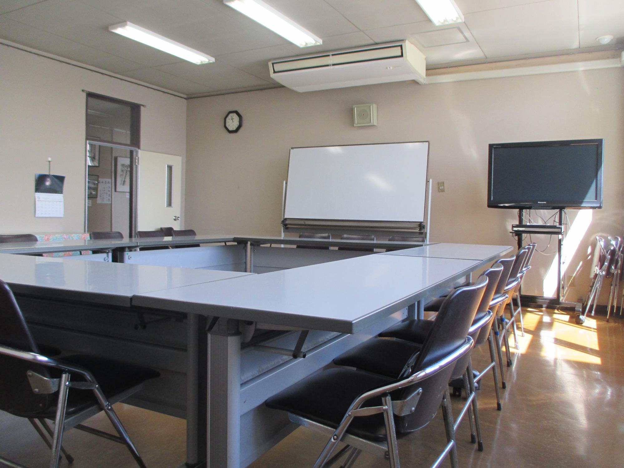 蛍光灯とエアコンを備えた白い天井、正面の白いホワイトボード、右側に壁掛けテレビ、左側に出入口があり、灰色の長机と黒い折りたたみ椅子が整然と配置された視聴覚室の写真