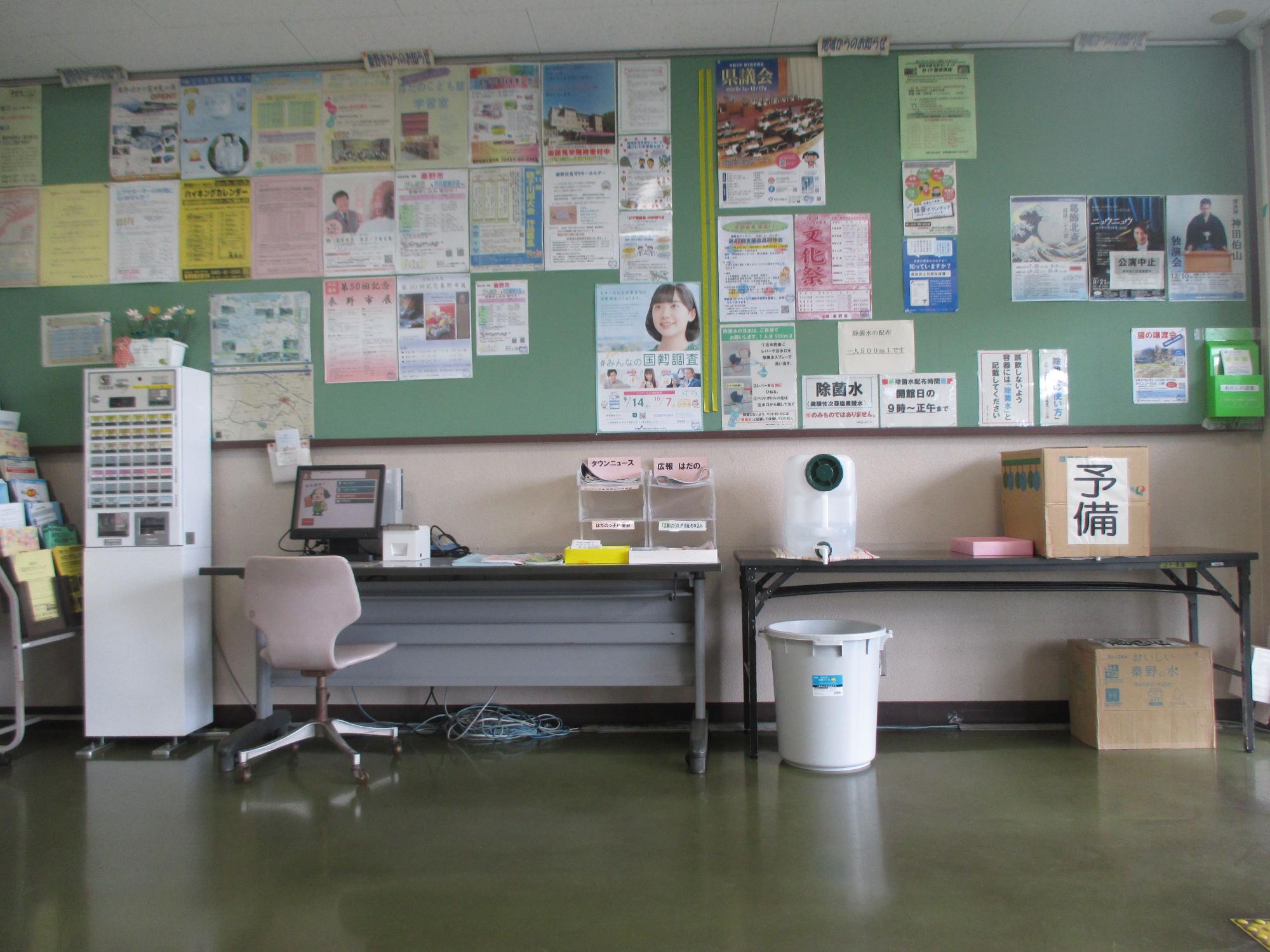 緑色の掲示板に多数のポスターやお知らせが掲示され、下部には白い椅子のある机とコンピューター、加湿器、右端に「予備」と書かれた段ボール箱などが配置された公民館のロビー付近の写真