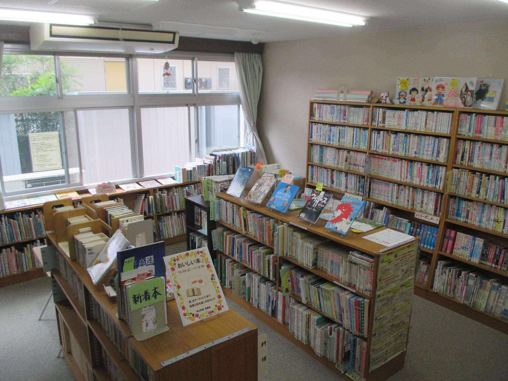 大きな窓から自然光が入る明るい図書室で、木製の本棚に多数の書籍と漫画が収納され、手前の展示台には新着本や推薦図書が並び、背後の高い書棚の上部にはアニメキャラクターの装飾が飾られている写真