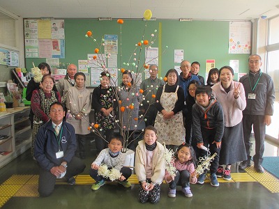 薄い緑色の壁に沢山の掲示物が貼られた室内で、高齢者から子どもまで幅広い世代の人々が、カラフルな餅花の飾り付けがされた木を囲んで記念撮影をしている様子を写した写真