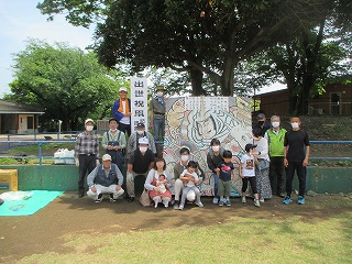 大きな木が立っている広場に老若男女が集まり、出世祝と書かれた看板と、武士の絵が描かれた大きな絵を背景に、家族連れや年配者が笑顔でポーズをとっている集合写真