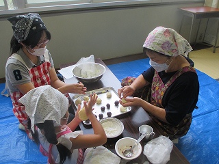 畳の上にブルーシートが敷かれた室内で、エプロンとバンダナを身につけた2人の女性と女の子が長机を囲み、楽しそうに団子作りをしている様子を捉えた写真