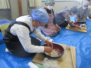 エプロンと三角巾を身につけた男女複数人が青いビニールシートの上で、ボウルの中のそば粉を混ぜたり麺棒で伸ばしたりするそば打ち体験の様子を捉えた写真