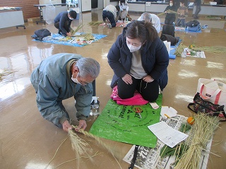 床にビニールシートを敷いた場所で間隔をあけて他の参加者が藁を使った作業をしている室内で、年配の男性が藁を編んでおり、隣に座った女性がその作業を正座をして見守っている様子を捉えた写真