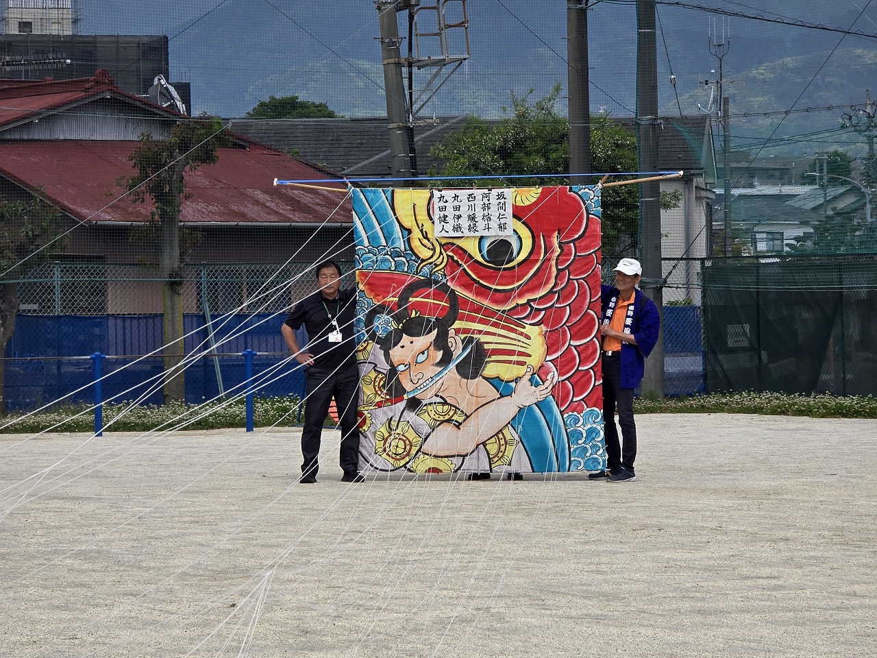 和柄のイラストが描かれた大きな凧を、地面に広げて二人で持ち、凧揚げの準備をしている様子の写真